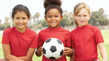 Three kids in red shirts on a field. The middle child is holding a soccer ball.