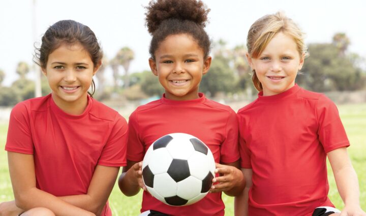 Three kids in red shirts on a field. The middle child is holding a soccer ball.