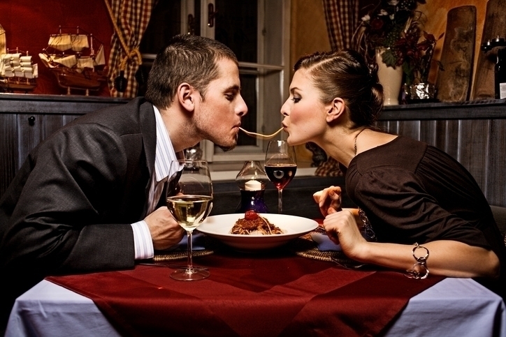 Couple dressed up and sharing a single strand of pasta at a romantic dinner.