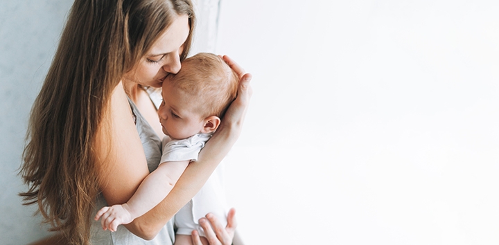 Woman holding a baby and kissing the baby on the head in front of a white background.