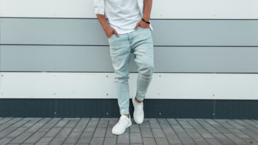 Young stylish man in a white fashionable t-shirt in vintage blue jeans in white trendy sneakers stands near a modern wall