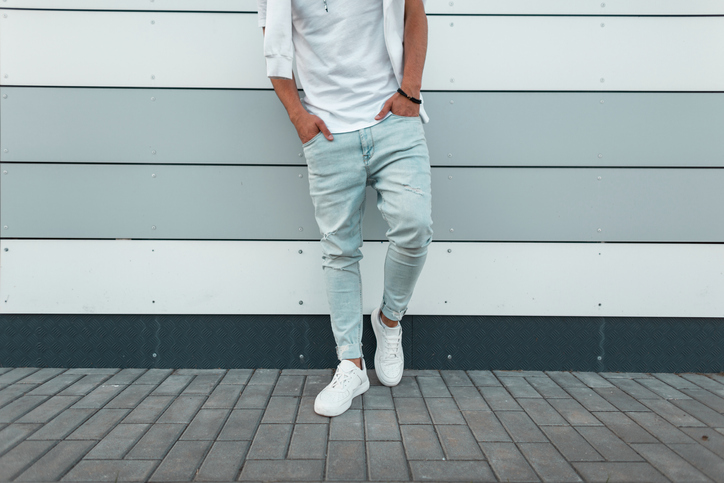 Young stylish man in a white fashionable t-shirt in vintage blue jeans in white trendy sneakers stands near a modern wall