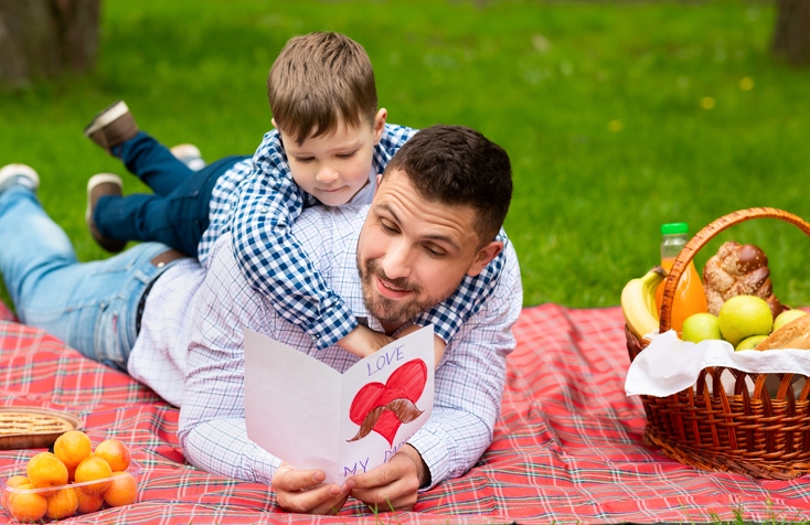 Cheerful dad reading his sons greeting card on picnic in public park.