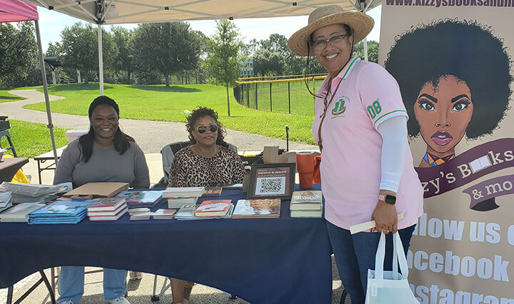 Two women sitting behind a table with books. One woman standing in front of the table.