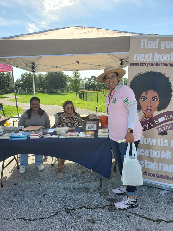 Two women sitting behind a table with books. One woman standing in front of the table.