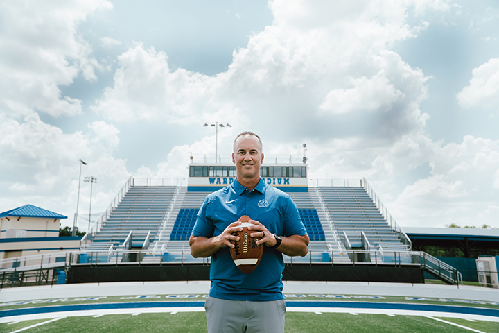 Coach Jeff Conaway holding a football, standing in the middle of the TFA football field.