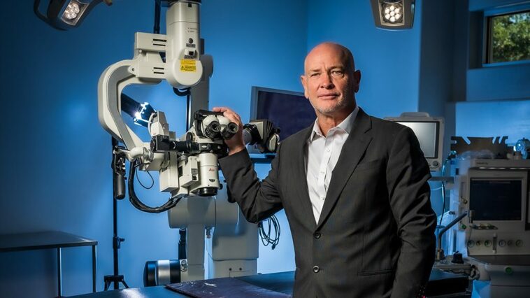 Dr. Masson wearing a black suit, standing next to medical equipment in a blue operation room.