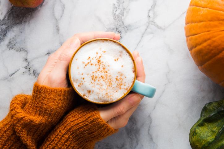 Autumn moody background with mug of pumpkin spice latte coffee and pumpkins on marble table. Flat lay in fall colors. Female hands in cozy sweater