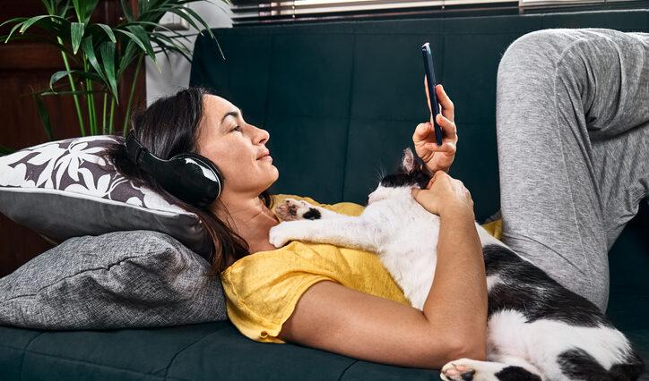 Woman listening to a true crime podcast with headphones lying embraced with her cat on the sofa.