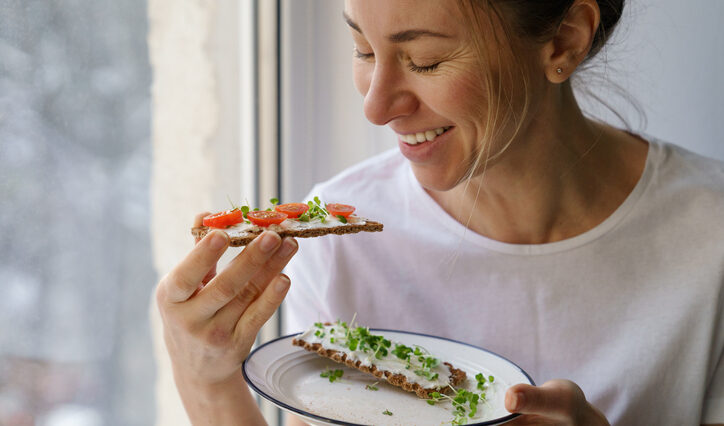 Smiling woman eating rye crisp bread with creamy vegetarian cheese tofu, cherry tomato and rucola micro greens, sitting at home and looking at window. Healthy food, gluten free, diet concept.