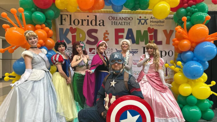 Captain America kneels with his shield, surrounded by Disney princesses under a rainbow balloon arch at a kids' event.